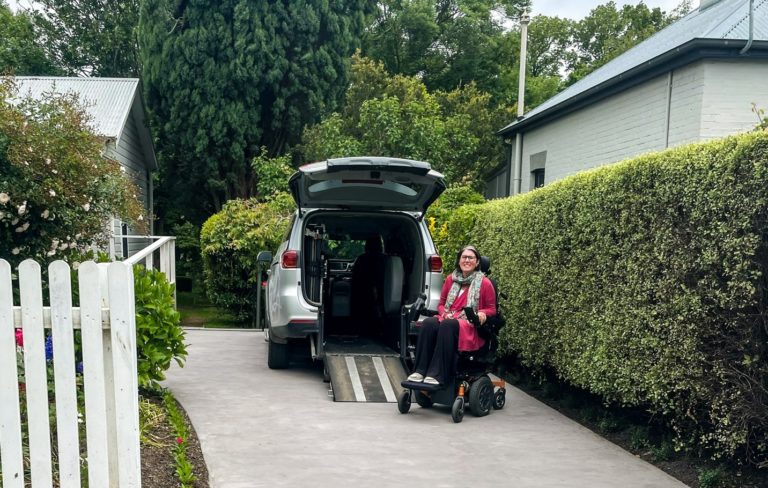 A young woman in a motorised wheelchair, on her driveway, showing how easily she can access her car