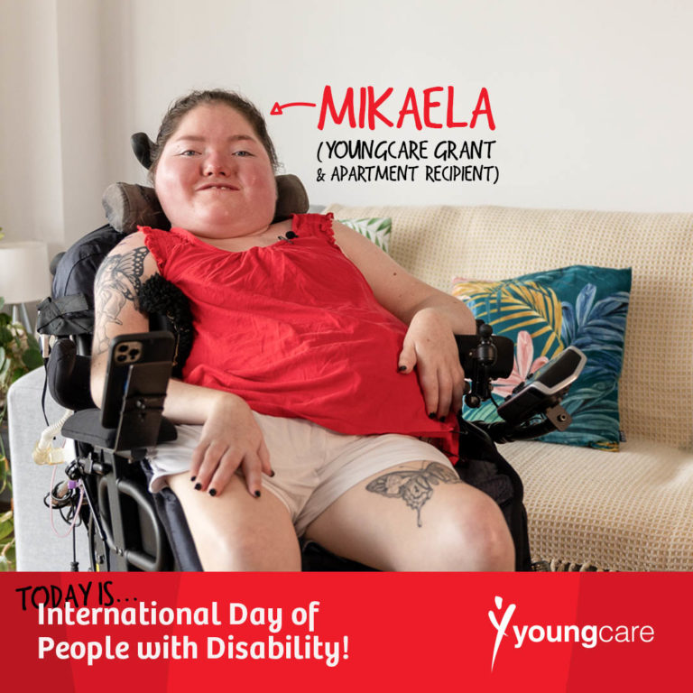 A young lady is smiling, sitting on an electric wheelchair inside an apartment's lounge room. She is wearing shorts and a red top. The room is light-filled and decorated with furniture and plants.