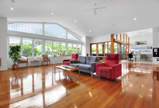 A well-lit living room with wooden floors, a mix of sofas, and large windows overlooking greenery, extending to an office and kitchen area.