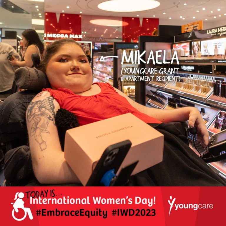 Mikaela is inside a modern makeup store, surrounded by displayed rows of beauty products, tall mirrors and star-like lights on the roof. Mikaela is wearing makeup from the store and is in a red top and black pants. They're in an electric wheelchair and have a makeup box on their lap. Words are positioned beside them with an arrow pointing in their direction reading "Mikaela (Youngcare Grant & Apartment Recipient")". A banner under the image reads "Today is International Women's Day! #EmbraceEquity #IWD2023". There is a white Youngcare logo beside this text.
