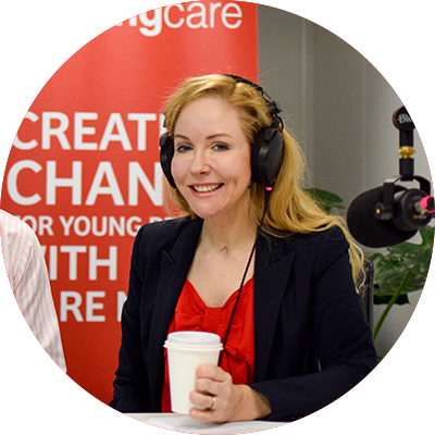 Headshot of Lisa Cox, an award-winning writer, presenter, and disability advocate. Lisa is wearing a red top, and black suit jacket. She's sitting in front of a podcast microphone, and is wearing recording headphones. She also has a takeaway coffee cup in her left hand. 