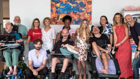 Group photo inside the lounge room of a Youngcare share house featuring three residents in electric wheelchairs, Margot Robbie, carers, family members and Youngcare team members.