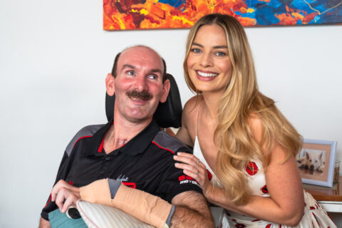 Margot Robbie and Carl are smiling for a group photo. Margot Robbie is wearing a white dress, and Carl, in an electric wheelchair, is wearing a black polo shirt.