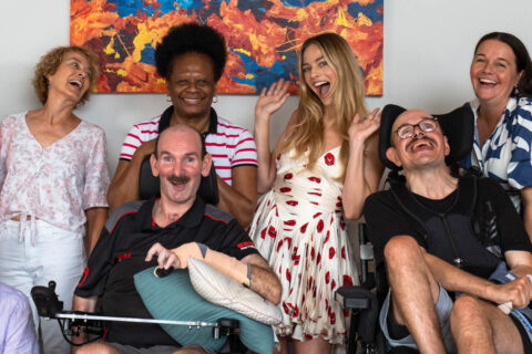 A fun group photo in a Youngcare share house. Two residents in electric wheelchairs are smiling joyfully, alongside Margot Robbie, carers and family members also smiling. Margot has her hands up in the air in celebration.