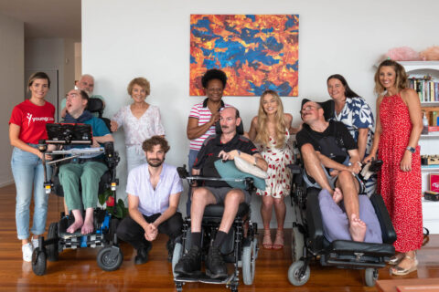 Group photo inside the lounge room of a Youngcare share house featuring three residents in electric wheelchairs, Margot Robbie, carers, family members and Youngcare team members.