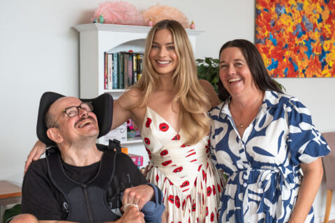 Margot Robbie, Brian and his sister, Tash, are smiling for a group photo. Margot Robbie is wearing a white dress. Brian, in an electric wheelchair, is wearing a black tee shirt. Tash is wearing a blue and white dress.